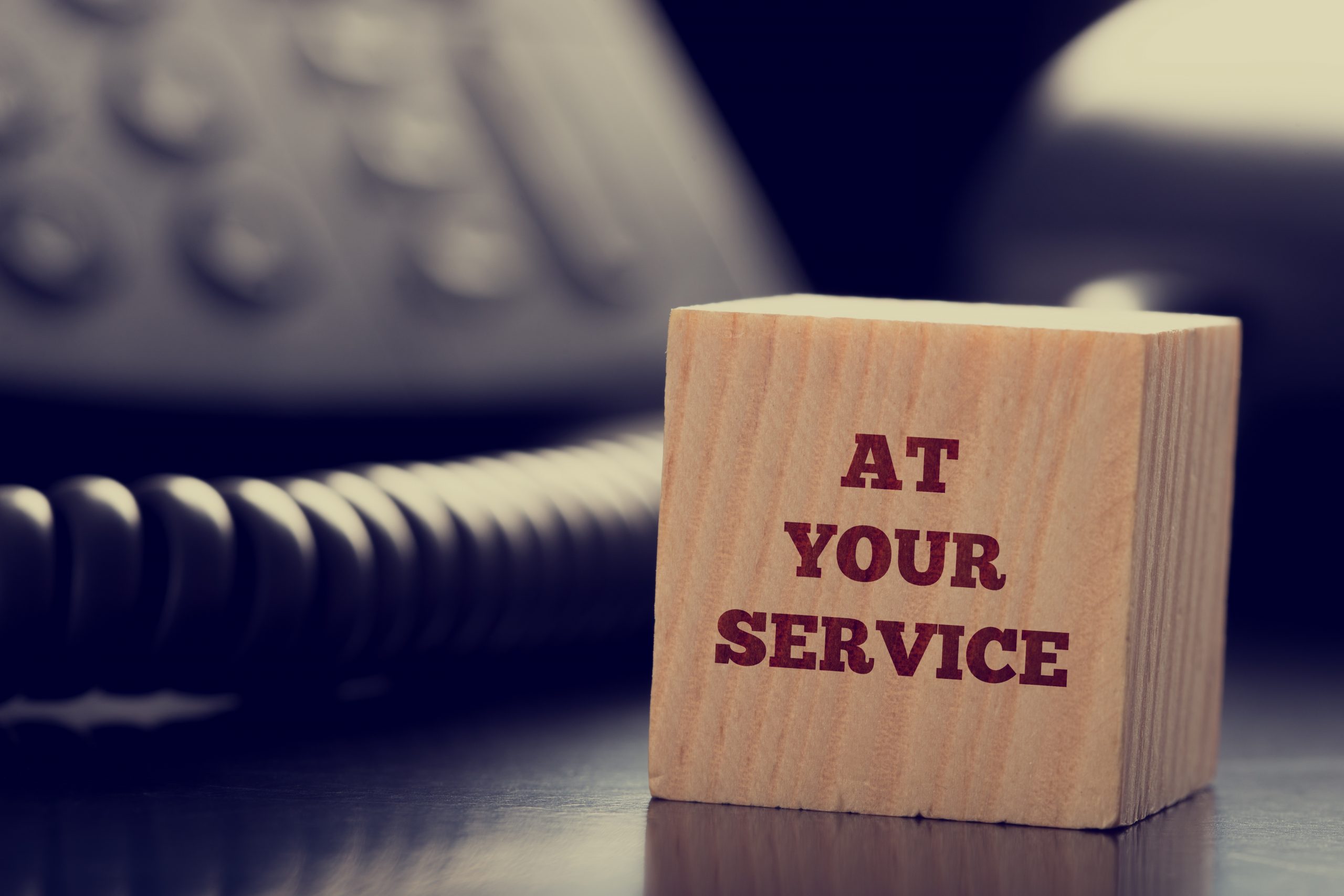 At Your Service written on a wooden cube in front of a telephone conceptual of help, client services, assistance, expertise and consultancy.